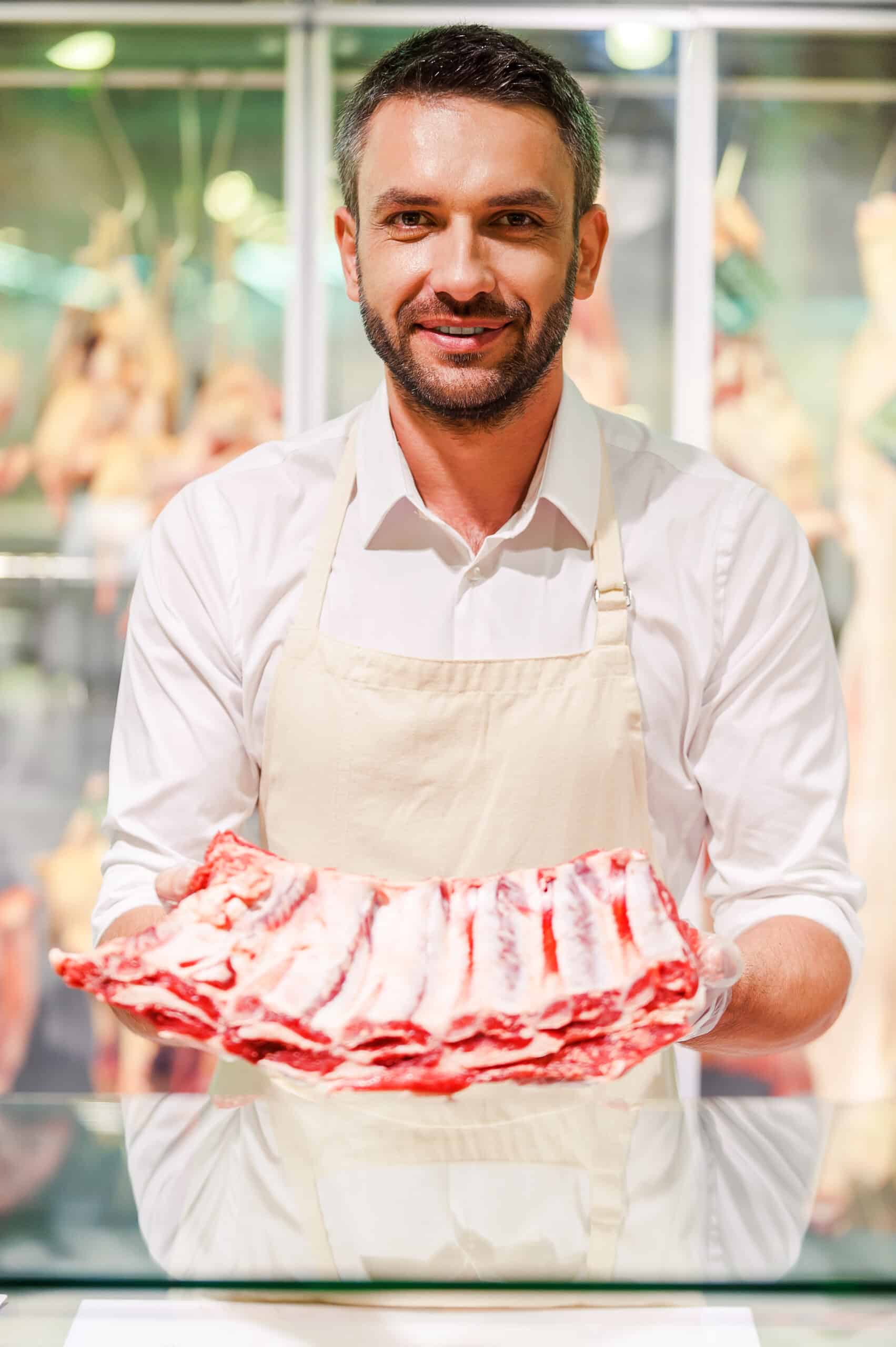 Only the freshest meat! Smiling young butcher stretching out slice of meat and looking at camera while standing at supermarket checkout